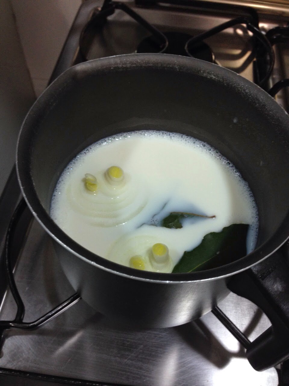 Leite fervendo em uma panela de metal com pedaços de cebola e uma folha de louro flutuando, tons de branco e creme