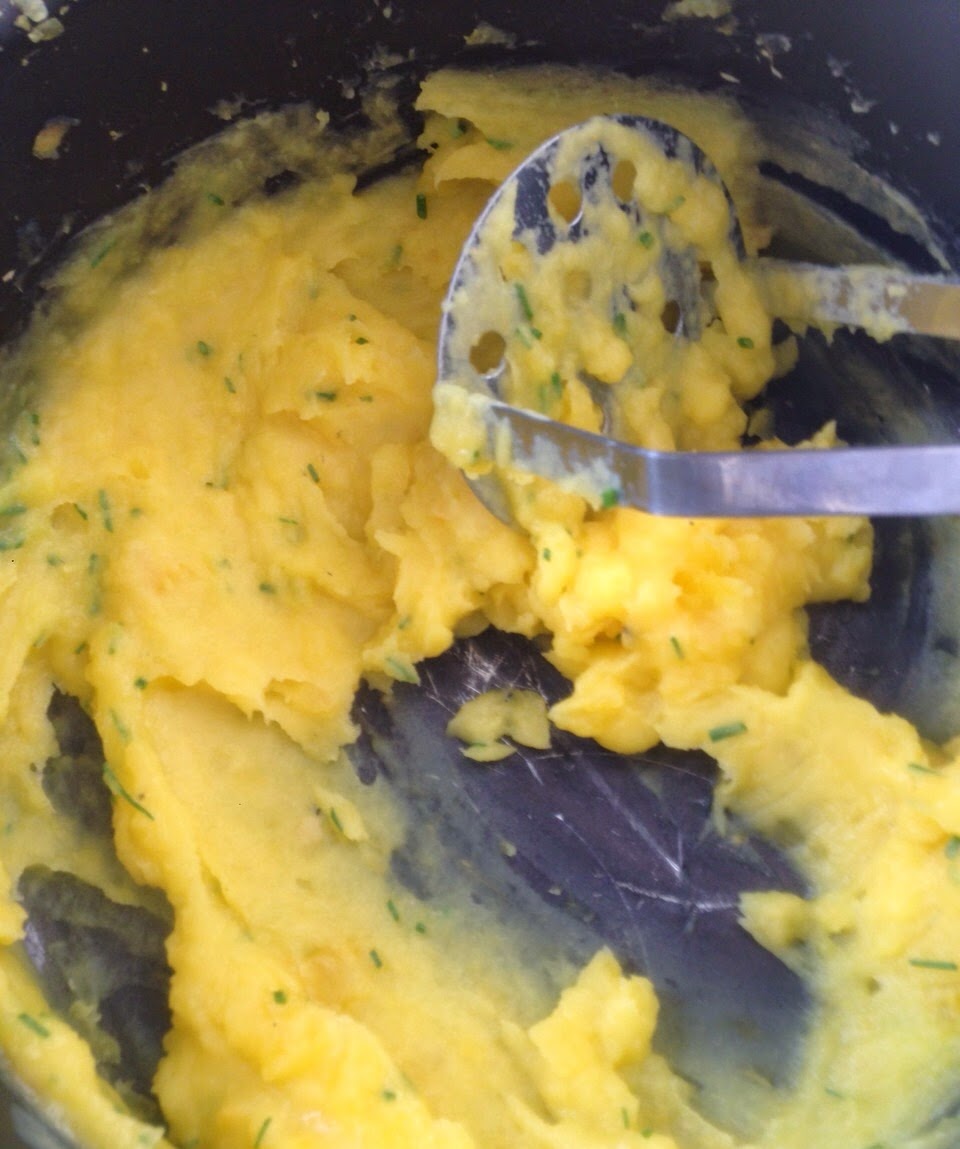 Purê de batatas com ervas picadas sendo amassado em panela escura, tons de amarelo e verde